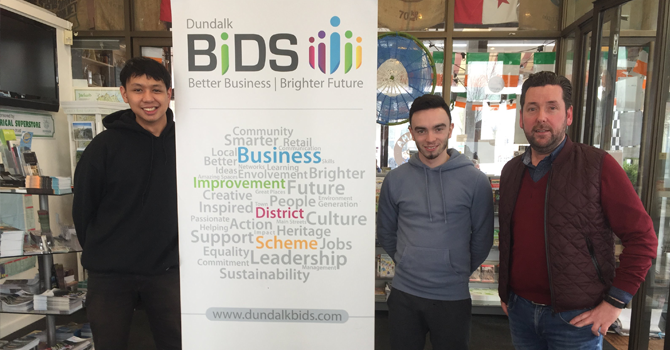 Three men standing beside a Dundalk Bids poster