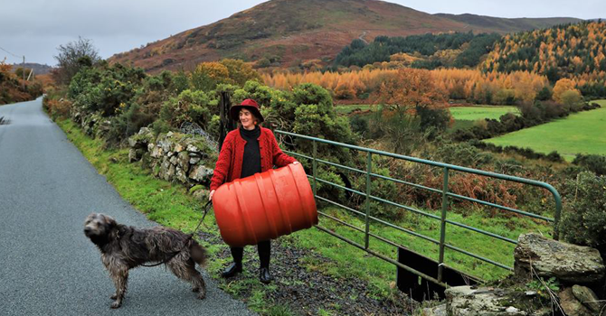 A woman in a red coat and hat stands on a country road with a dog, with a mountain in the background