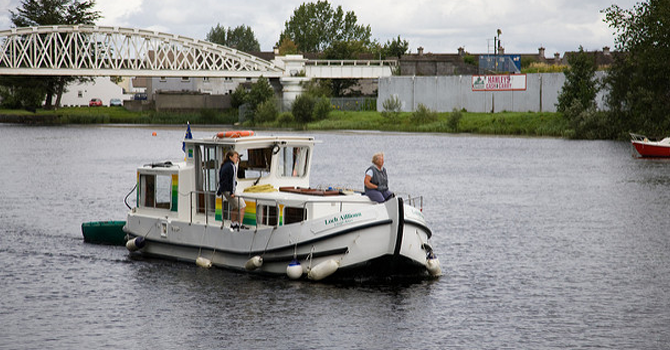 People sit on a small ferry on the River Shannon, with a bridge in the background.