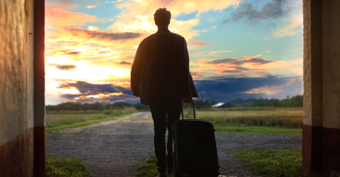 Image of a person's back walking away from camera, into the sunset and pulling a small suitcase