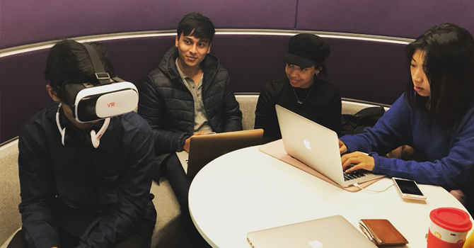 Students sit around a table. One has a VR headset on.