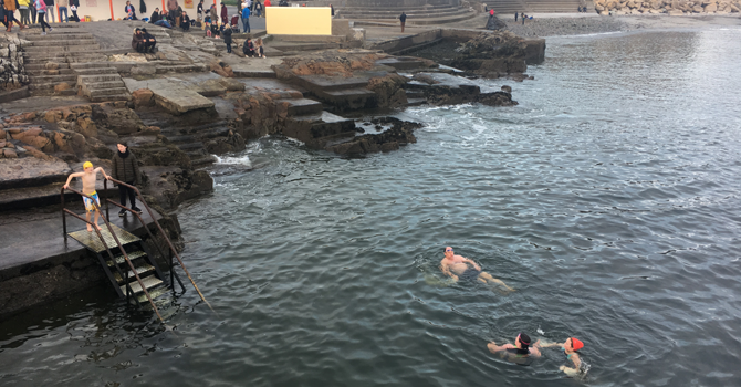 Three people swim in the sea while a boy descends a metal staircase into the water