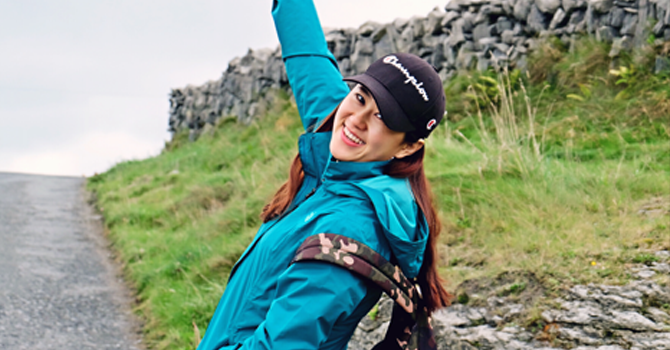 A girl with a cap and green coat smiles and poses for camera while on a walk on a grassy hillside