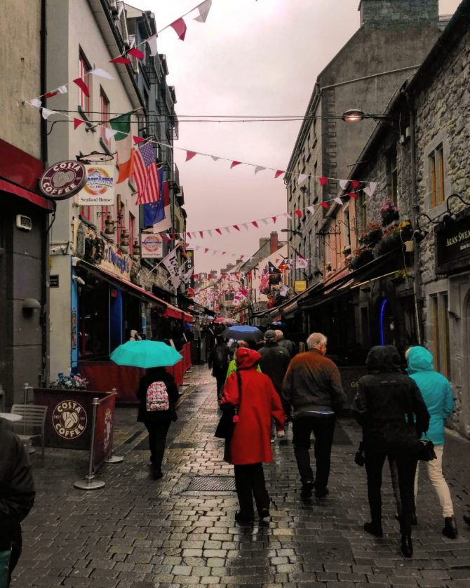 Peoplpe walking with umbrellas on a rainy cobbled side street as bunting flaps overhead 