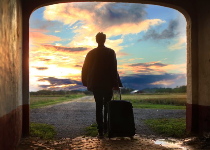 Man standing in archway with a suitcase watching the sun set 