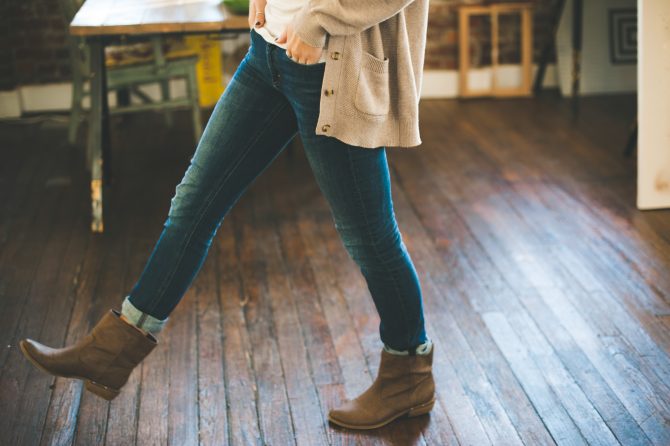Woman in beige boots blue jeans and a cream coloured cardigan with her foot raised as she walks across a wooden floor 