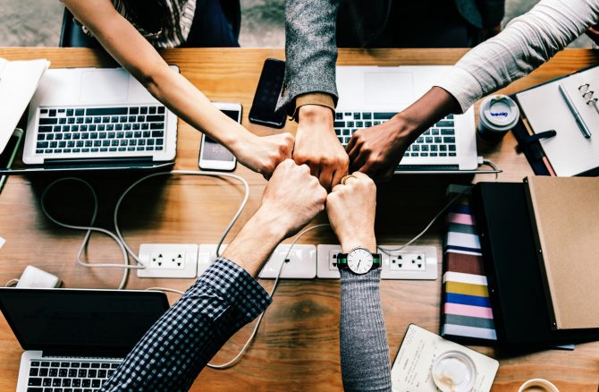 group of hands fist bumping over a desk containing computers and a power outlet 