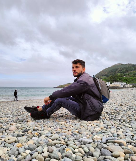 An image of a person sitting on a pebbled beach and facing the sea